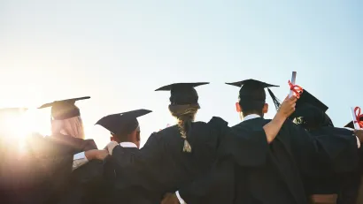 View from back of group of college students in caps and gowns at graduation with bright sunlight in front of them.